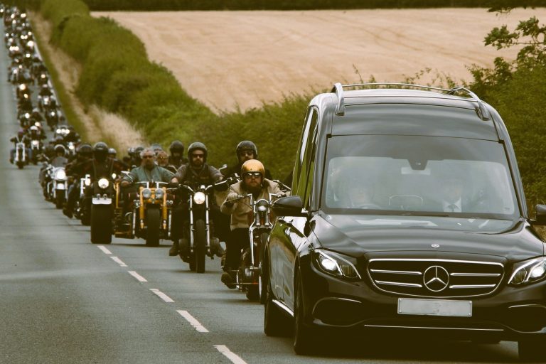 Black hearse driving along a road with hundreds of motorbikes following in a procession.