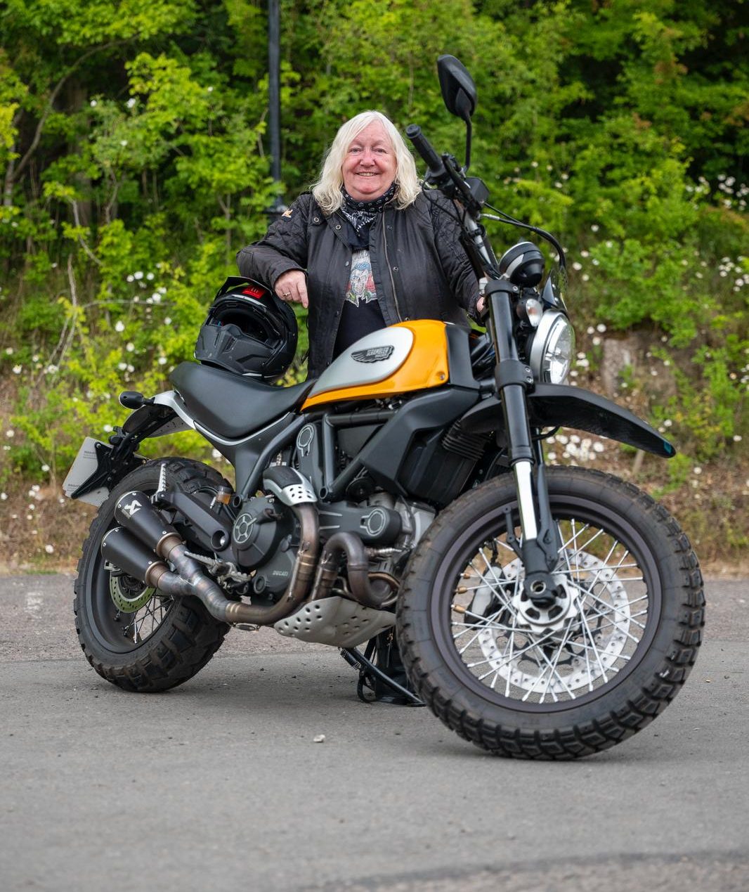 Woman with shoulder-length platinum-blonde hair leaning against her yellow and black Scrambler motorbike, wearing a biker jacket and jeans.